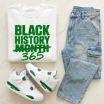 White t-shirt with 'Black History Month 365' text, white sneakers with green accents, and blue jeans on a light background