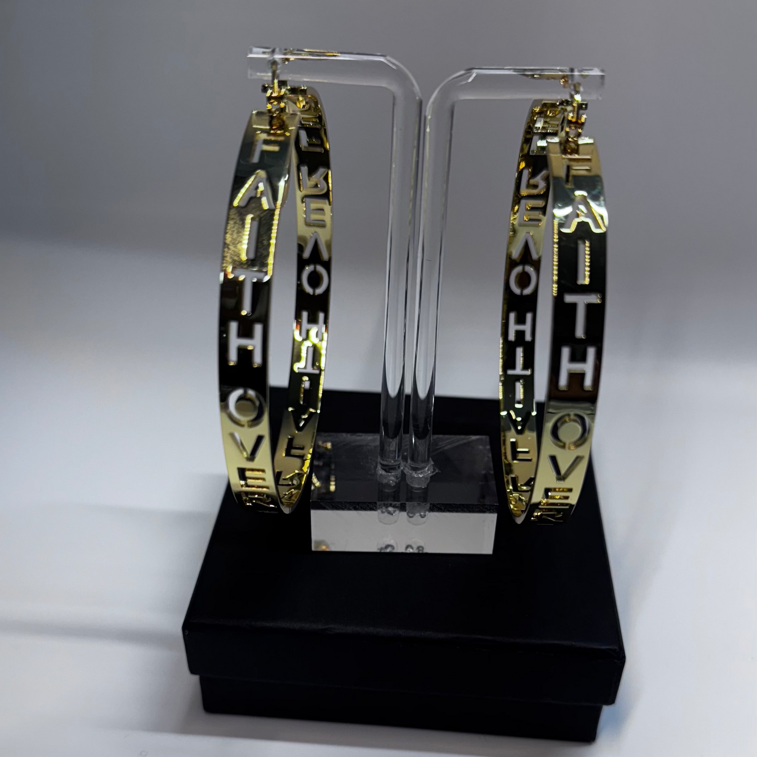 Hoop earrings with FAITH OVER FEAR GOLD EARRINGS' text on a clear stand against a gray background