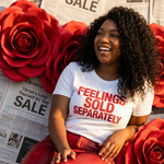 Woman sitting on a couch with red flowers and newspaper in the background wearing a white t-shirt with red text.