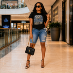 A woman stands in a mall corridor wearing a black graphic T-shirt that reads 'GOD CAN'T PLAY ABOUT ME' in distressed blue and white lettering, paired with light-wash denim cutoff shorts, black strappy heels, and a black quilted handbag with a gold YSL monogram.