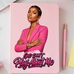 Notebook with a cover featuring a woman in a pink blazer and text 'God Don't Play About Me' on a desk with office supplies.