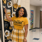 A woman stands in a school hallway wearing a yellow graphic T-shirt that reads 'I'M YOUR TEACHER thank you school' and a matching yellow-and-white plaid flared skirt. She is holding books and posing in front of a decorated teacher door display.