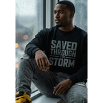 Person wearing a black sweatshirt with 'SAVED THROUGH THE STORM' text, sitting by a window with rain outside.