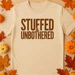 Beige t-shirt with 'STUFFED & UNBOtherED' text on a wooden surface with autumn leaves and pumpkins.