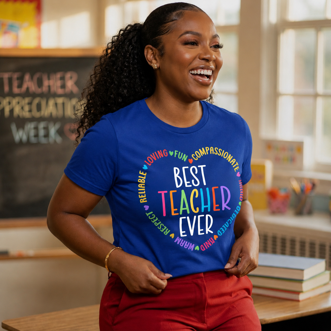 A woman smiling in a classroom while wearing a royal blue graphic T-shirt that reads 'BEST TEACHER EVER' in multicolored letters, paired with red pants.