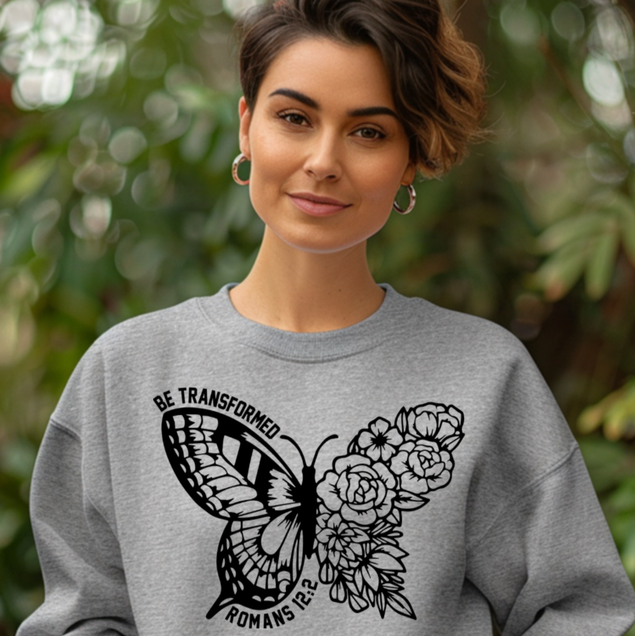 Person wearing a gray sweatshirt with a butterfly design and text "BE_TRANSFORMED"outdoors.