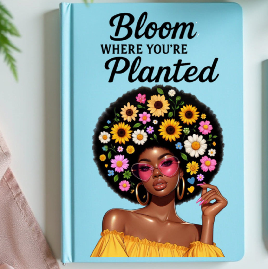 JOURNAL with a design of a woman with flowers in her hair and 'Bloom Where You're Planted' text, placed on a surface with a cup of coffee and plant.