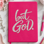 A hot pink hardcover journal with the phrase 'but GOD.' in glittery silver script on the cover, styled on a white marble surface with pink roses, a cross, a pen, and a coffee cup nearby.
