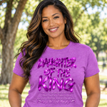 A woman wearing a purple graphic t-shirt with 'DAUGHTER OF THE KING' printed in metallic pink letters, standing outdoors in front of a blurred green background.