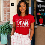 A woman wearing a bright red graphic t-shirt with the text 'DEAN guide encourage inspire' and white plaid pants, standing in front of a school doorway.