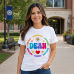 A woman stands outdoors on a campus walkway wearing a white graphic T-shirt with a colorful 'DEAN' design and blue jeans. The background shows a brick building, trees, a bench, and flowers.