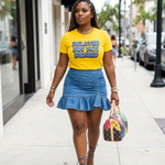 Woman walking on a sidewalk wearing a yellow t-shirt with text "DELAYED BUT NOT DENIED" and a blue denim skirt. 