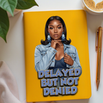 Yellow notebook with a portrait of a woman and 'Delayed But Not Denied' text, placed on a white surface with a plant and coffee cup in the background.