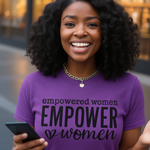 Woman wearing a purple t-shirt with 'empowered women EMPOWER women' text, holding a phone.