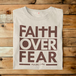 A folded off-white t-shirt with a brown graphic reading 'FAITH OVER FEAR' and 'PSALMS 118:6' printed on the front, laid flat on a rustic wooden plank background.