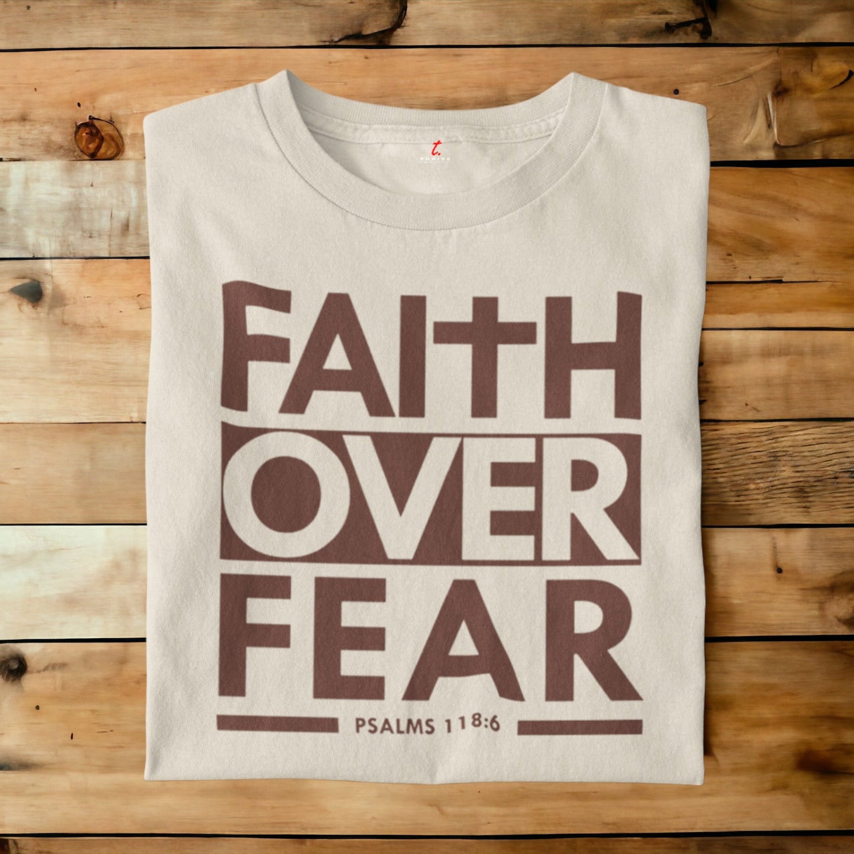 A folded off-white t-shirt with a brown graphic reading 'FAITH OVER FEAR' and 'PSALMS 118:6' printed on the front, laid flat on a rustic wooden plank background.