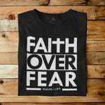 A black short-sleeve t-shirt with a bold white graphic reading 'FAITH OVER FEAR' and 'PSALMS 118:4' printed on the front, laid flat on a rustic wooden plank background.
