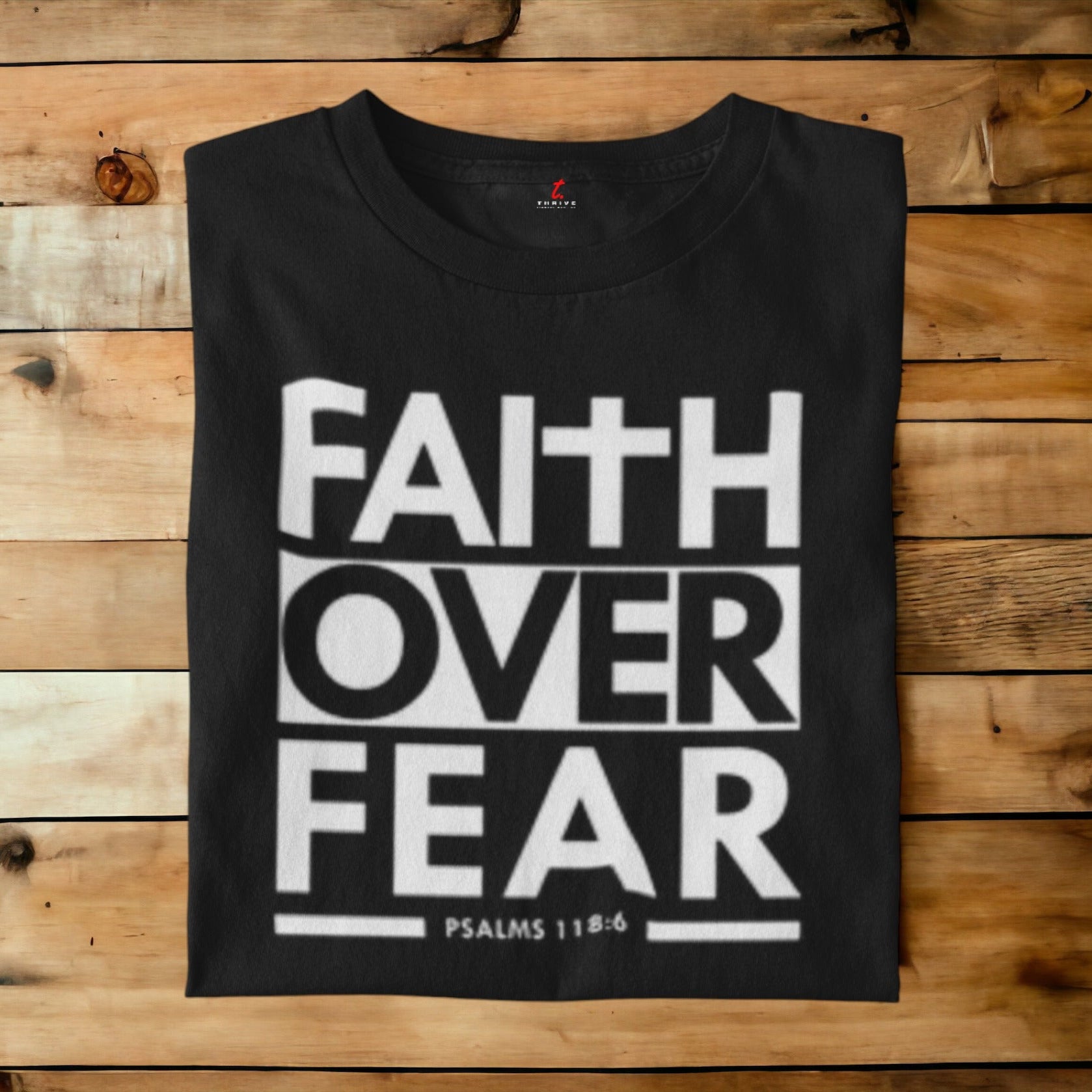 A black short-sleeve t-shirt with a bold white graphic reading 'FAITH OVER FEAR' and 'PSALMS 118:4' printed on the front, laid flat on a rustic wooden plank background.