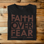 A black crewneck t-shirt with a brown graphic reading 'FAITH OVER FEAR' and 'PSALMS 118:6' laid flat on a rustic wooden plank background.