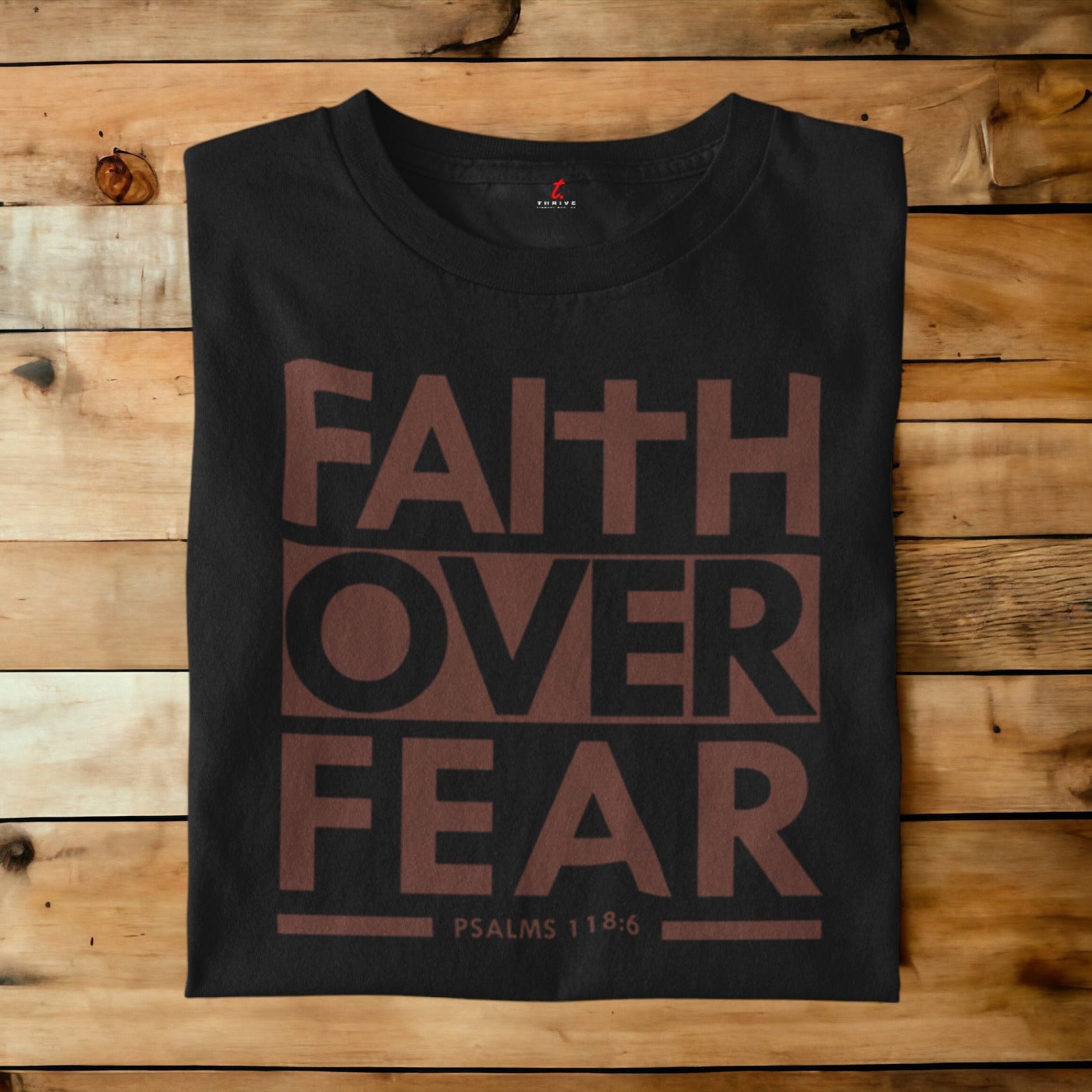 A black crewneck t-shirt with a brown graphic reading 'FAITH OVER FEAR' and 'PSALMS 118:6' laid flat on a rustic wooden plank background.