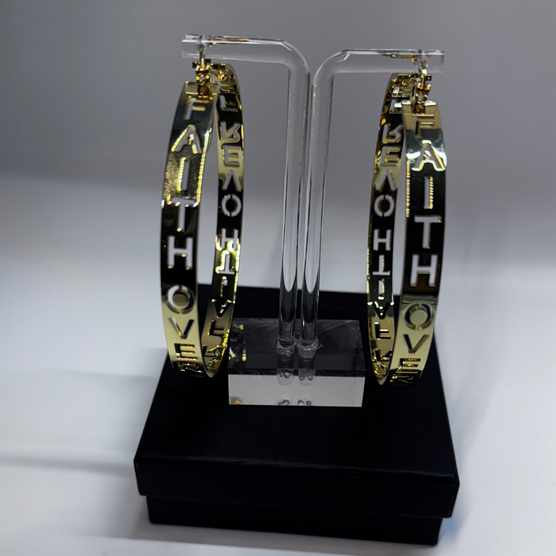 Hoop earrings with FAITH OVER FEAR GOLD EARRINGS' text on a clear stand against a gray background