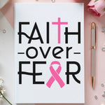 Notebook with 'Faith over Fear' text and pink ribbon on a decorative background with flowers and a pen.