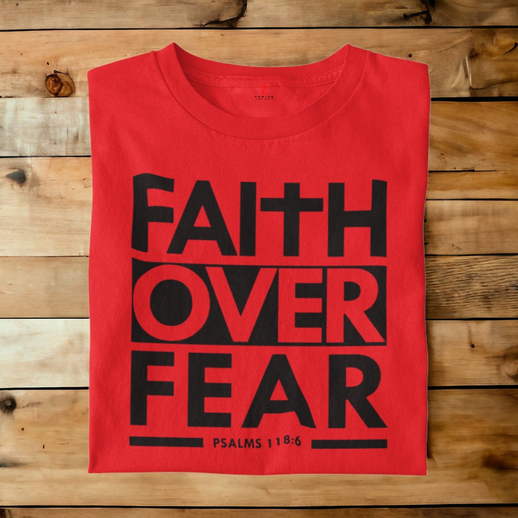 A red short-sleeve t-shirt with a bold black graphic reading 'FAITH OVER FEAR' and 'PSALMS 118:6' printed on the front, laid flat on a rustic wooden plank background.