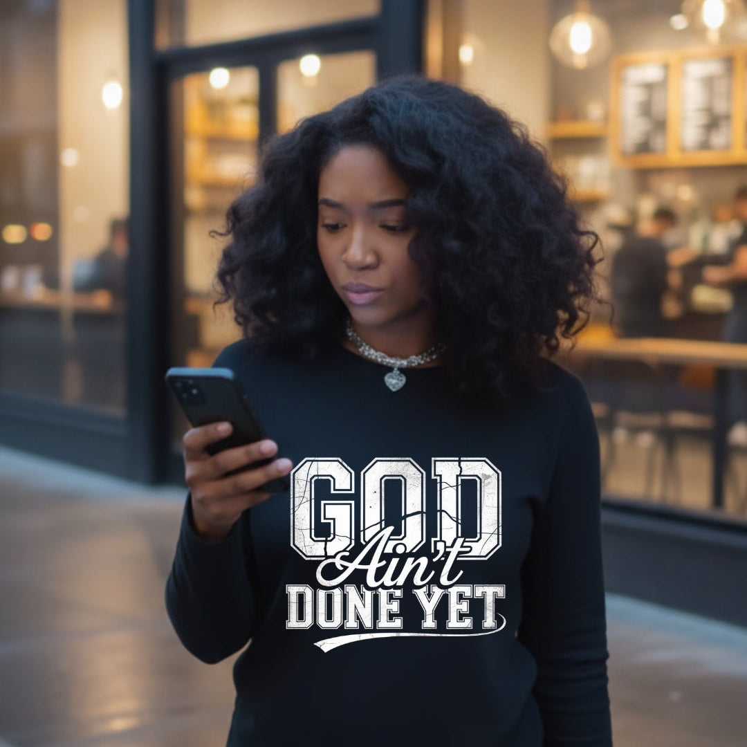 Woman wearing a black sweatshirt with 'God Ain't Done Yet' text, standing outside a building.