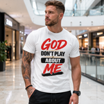 A man wearing a white graphic t-shirt with the text 'GOD DON'T PLAY ABOUT ME' in red and black, standing in a modern shopping mall.