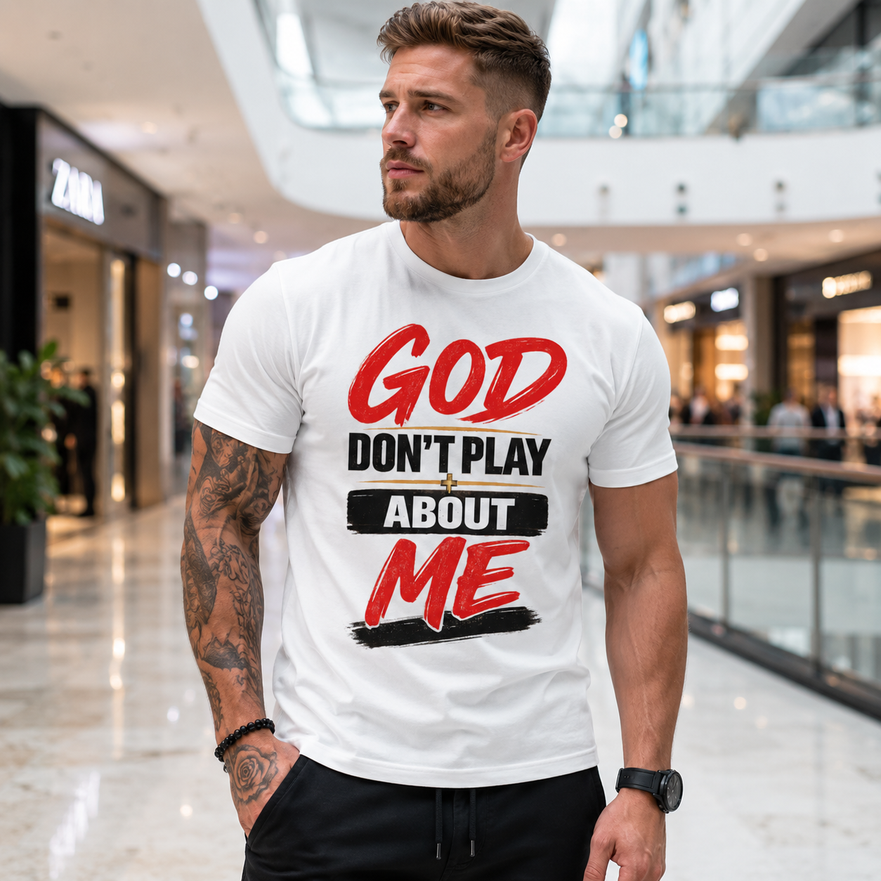A man wearing a white graphic t-shirt with the text 'GOD DON'T PLAY ABOUT ME' in red and black, standing in a modern shopping mall.