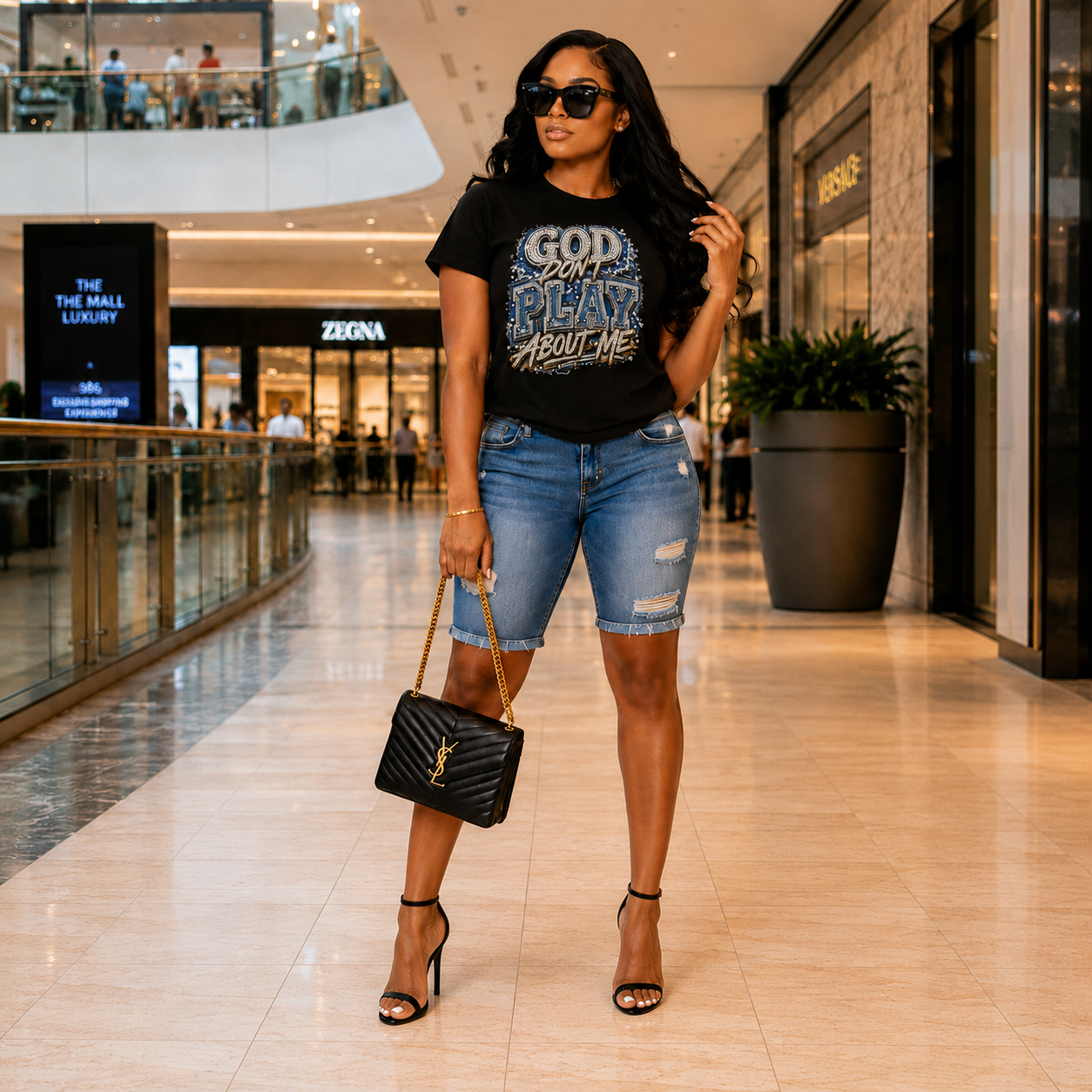 A woman stands in a mall corridor wearing a black graphic T-shirt that reads 'GOD CAN'T PLAY ABOUT ME' in distressed blue and white lettering, paired with light-wash denim cutoff shorts, black strappy heels, and a black quilted handbag with a gold YSL monogram.