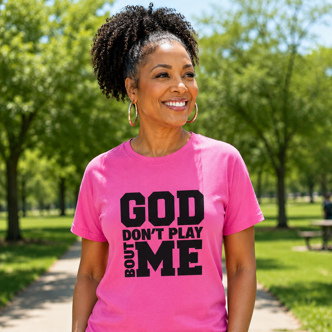 A smiling woman standing outdoors on a park path wearing a bright pink short-sleeve T-shirt with bold black text that reads 'GOD DON'T PLAY BOUT ME'.