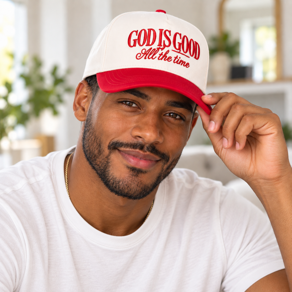 A man wearing a red and white trucker hat with the text 'GOD IS GOOD All the time' in red script, posing in a bright, modern indoor setting.