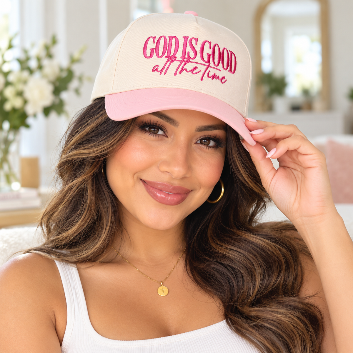 A woman wearing a beige and pink baseball cap with the embroidered text 'GOD IS GOOD all the time' in pink script, posing in a bright, softly lit indoor setting.