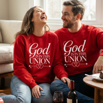 Two people wearing red sweatshirts with 'God is in this union' text, sitting on a couch in a living room.