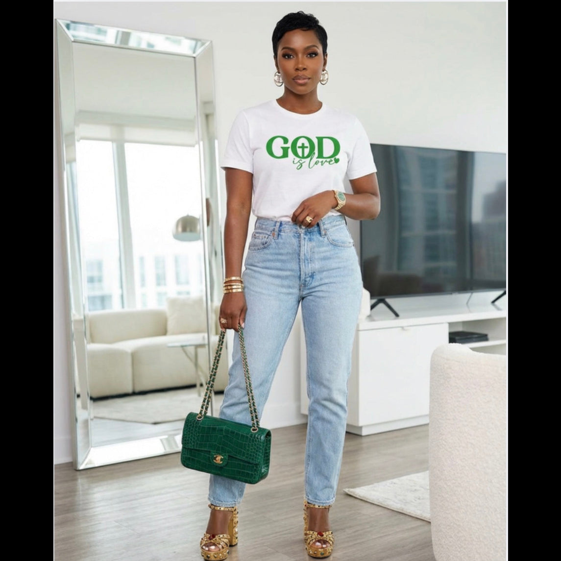 Woman wearing a white t-shirt with 'GOD is Love' text, blue jeans, and holding a green handbag in a modern living room.