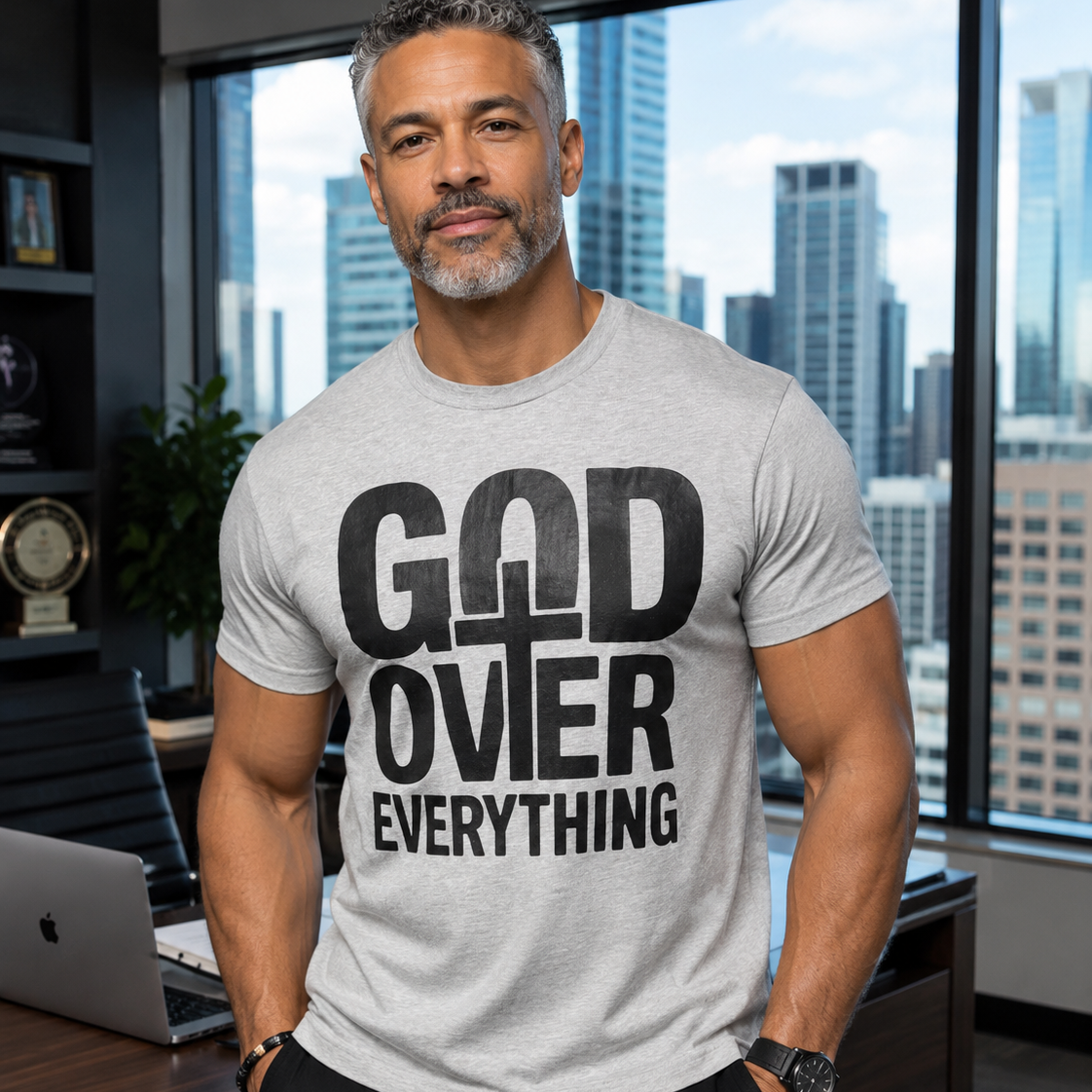 A muscular man with gray hair and a beard wearing a light gray t-shirt with a bold black graphic that reads 'GOD OVER EVERYTHING' with a cross symbol, standing in a modern office with a city skyline background.