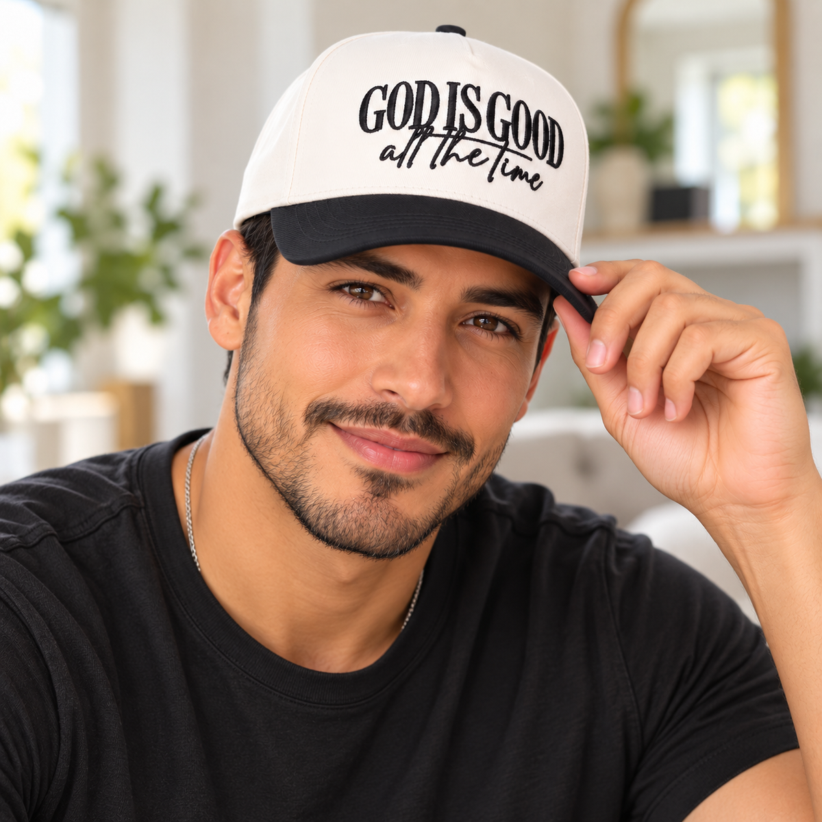 A man wearing a white and black snapback cap with the text 'GOD IS GOOD all the time' in black script, posing in a bright indoor setting.