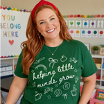 A smiling woman with red hair wearing a green graphic t-shirt that reads 'helping little minds grow' in white script, paired with blue jeans and a red headband in a colorful classroom setting.