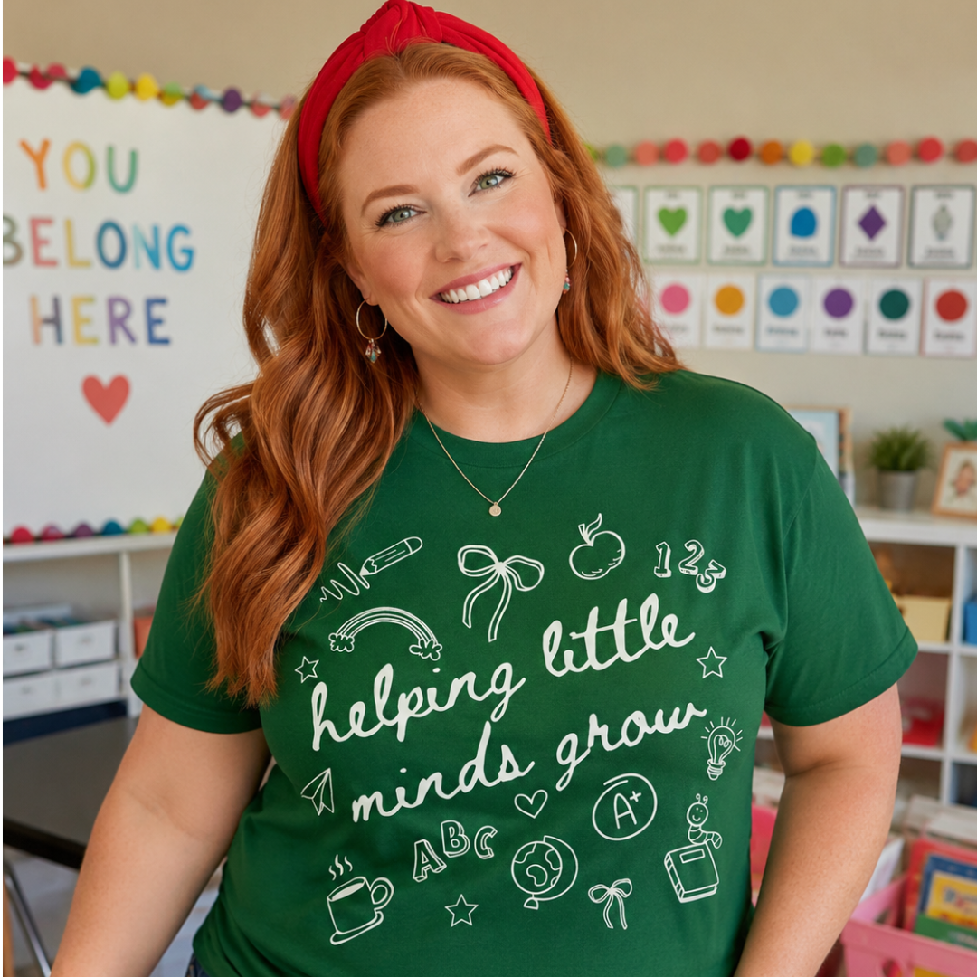 A smiling woman with red hair wearing a green graphic t-shirt that reads 'helping little minds grow' in white script, paired with blue jeans and a red headband in a colorful classroom setting.