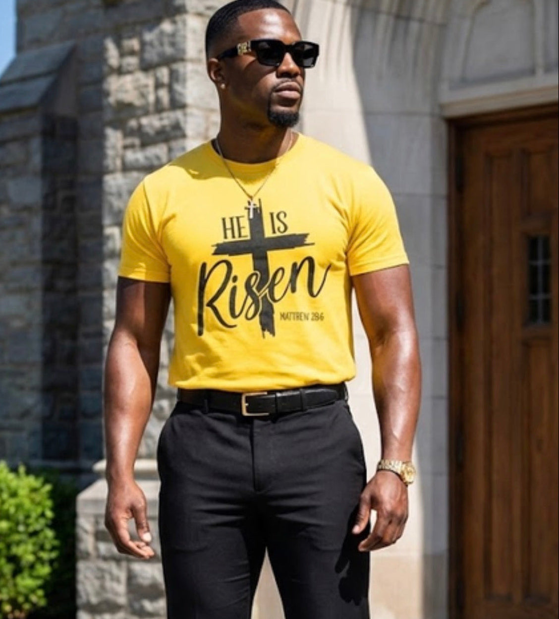 Man wearing a yellow t-shirt with 'He is Risen' text, standing in front of a stone building.