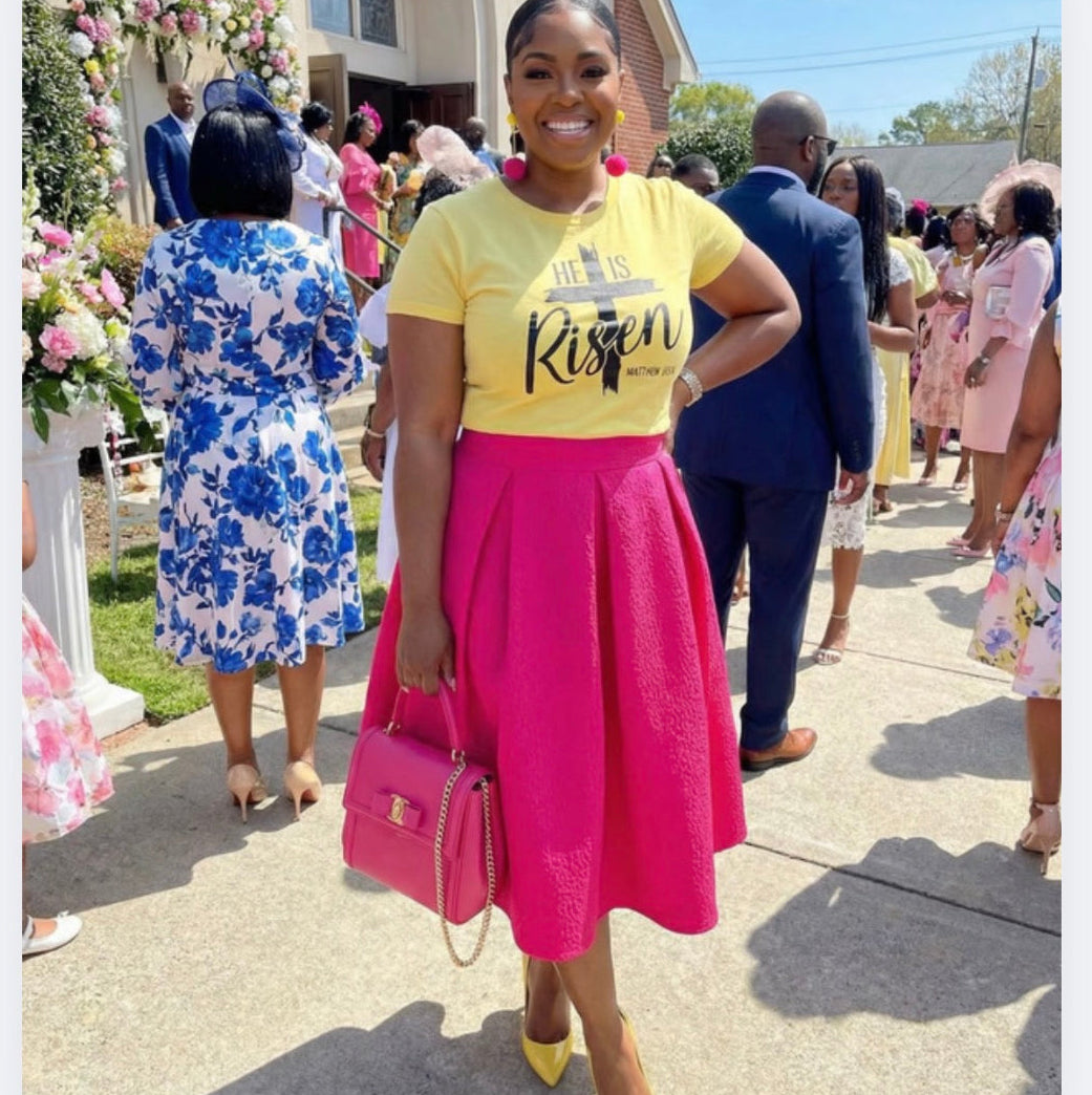 Woman in a yellow shirt with 'He is Risen' text, pink skirt, and yellow shoes standing outdoors with a church in the background.
