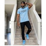 Man wearing a light blue t-shirt with text, black pants, and blue sneakers standing on a staircase.
