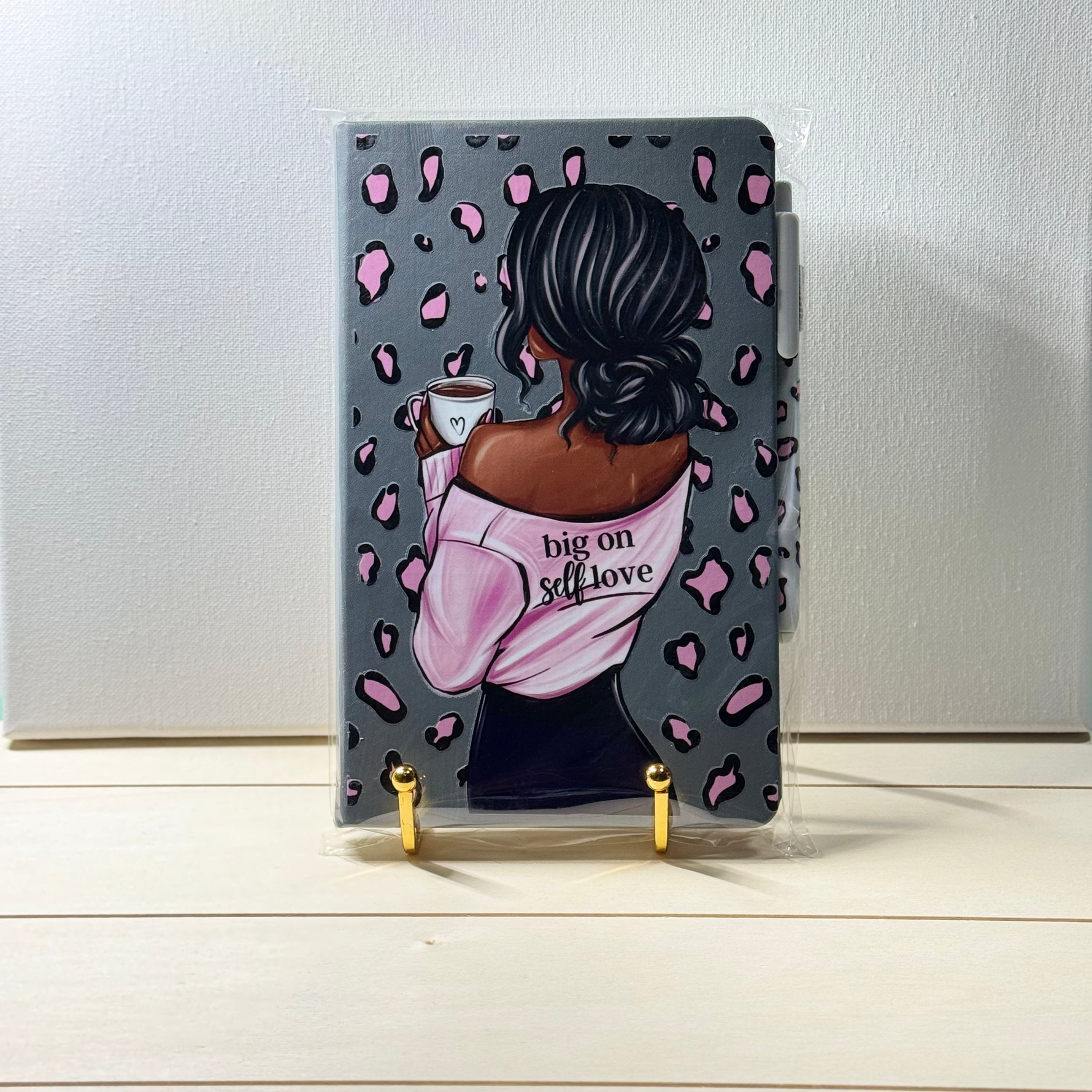Notebook with a design of a woman holding a coffee cup and text 'big on self love' on a white surface.
