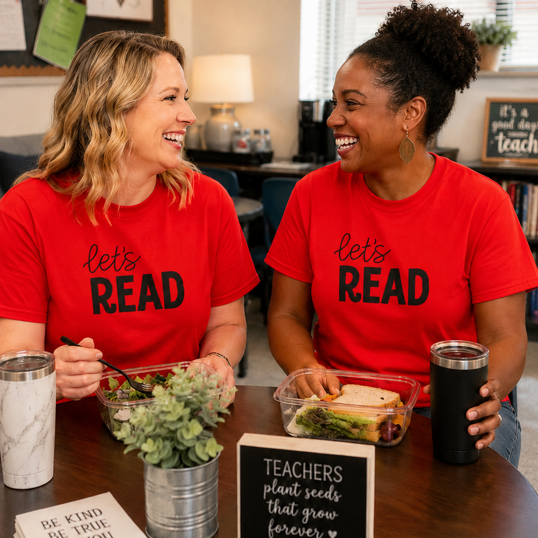 Two women sitting at a table wearing matching red graphic t-shirts that say 'let's READ' in black text. They are smiling and laughing while holding lunch containers and mugs in a bright classroom or office setting.