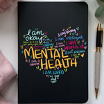 Black notebook with colorful mental health affirmations on a white surface with a pen and flowers.