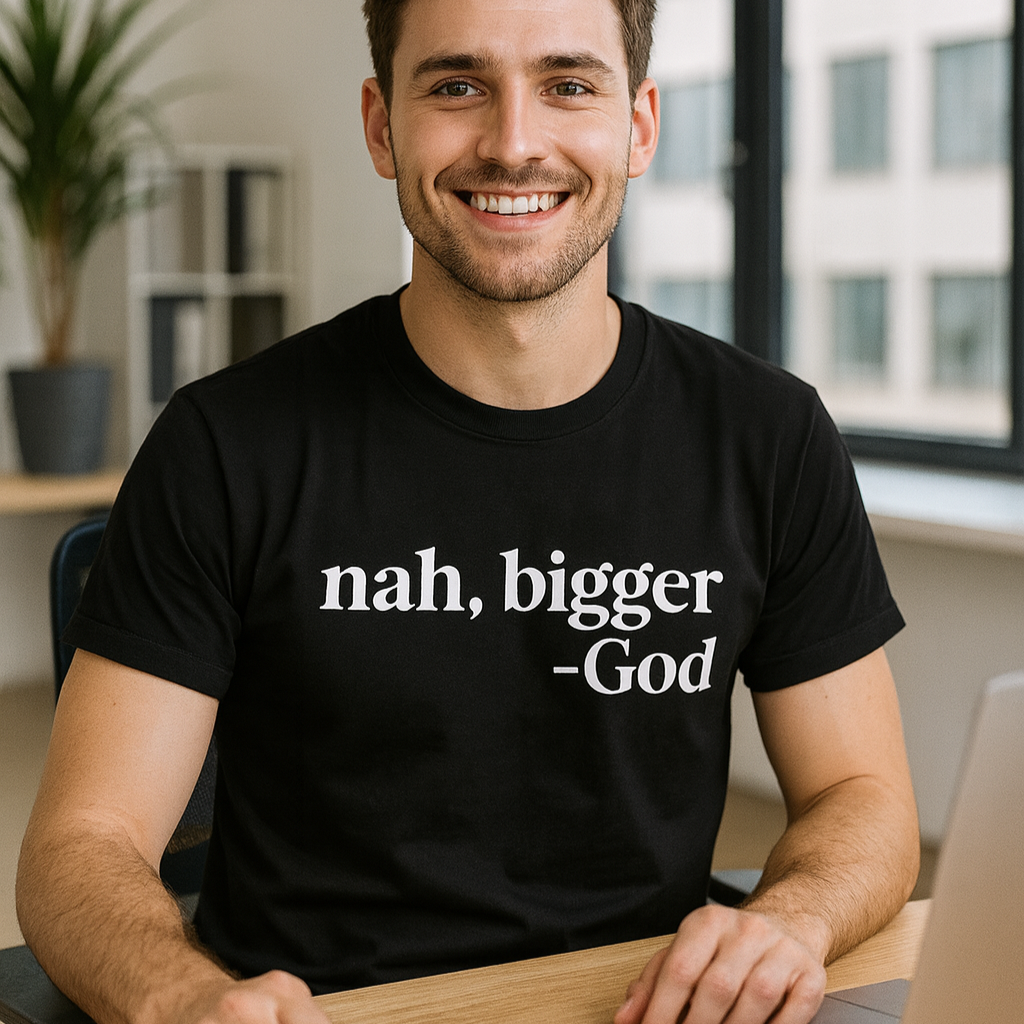 Man wearing a black t-shirt with 'nah, bigger -God' text, sitting at a desk with a laptop and notebook.