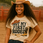 Woman wearing a beige cowboy hat and 'Not My First Rodeo' t-shirt in a dirt arena.
