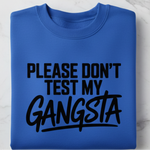 Blue sweatshirt with text 'PLEASE DON'T TEST MY GANGSTA' on a marble surface, with sunglasses and a plant in the background.
