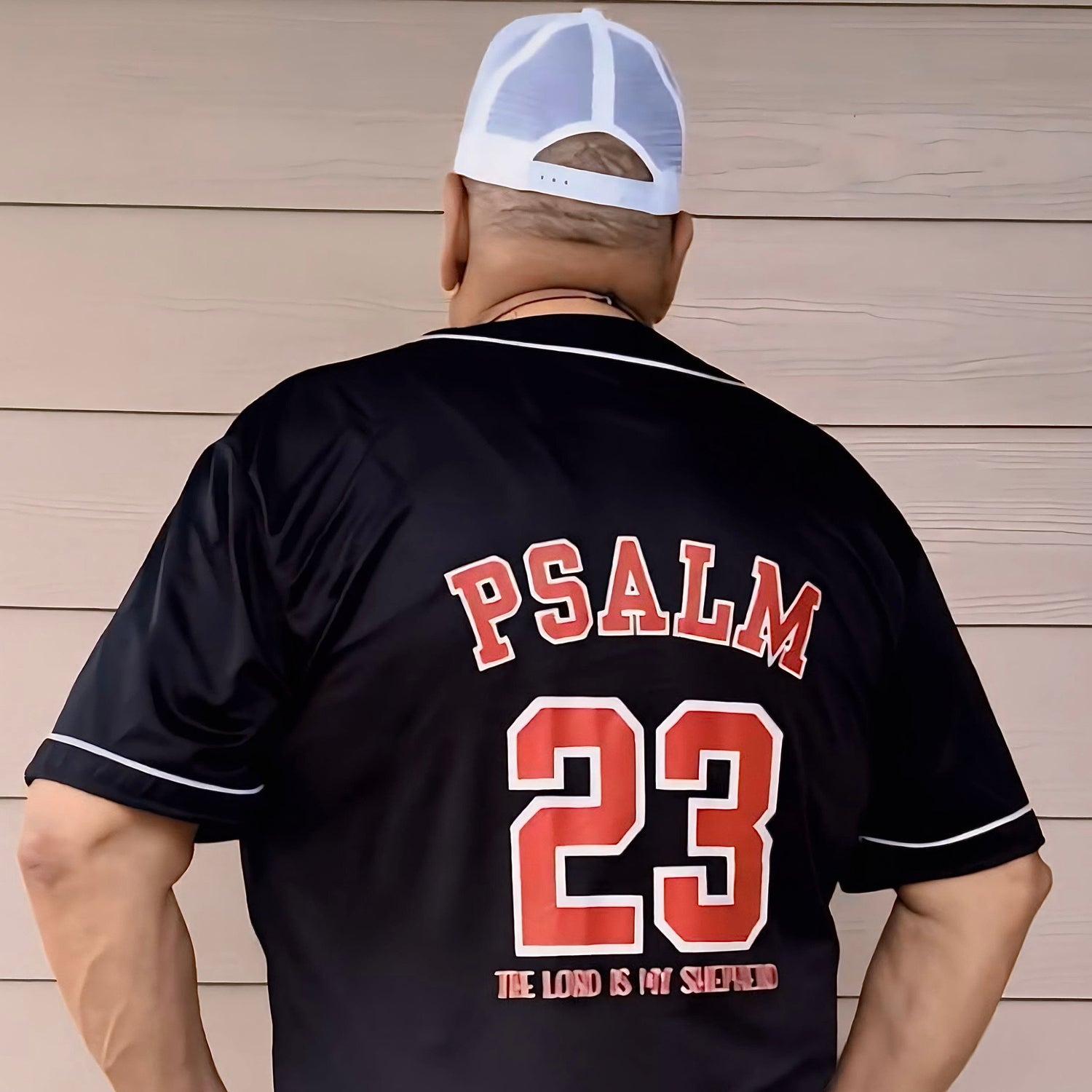 Person wearing a black baseball jersey with 'PSALM 23' printed on the back, standing against a wooden wall.
